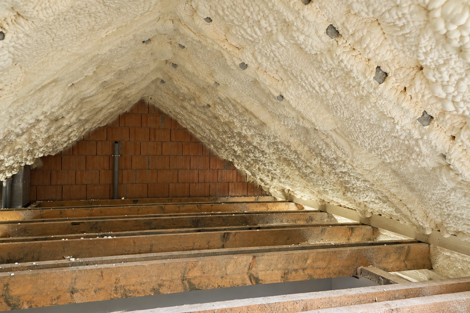 White spray foam coats the underside of an attic roof, with exposed wooden floor joists below.