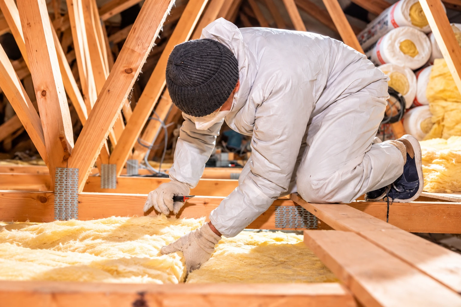 A worker in protective clothing lays yellow fiberglass batts between attic floor joists.
