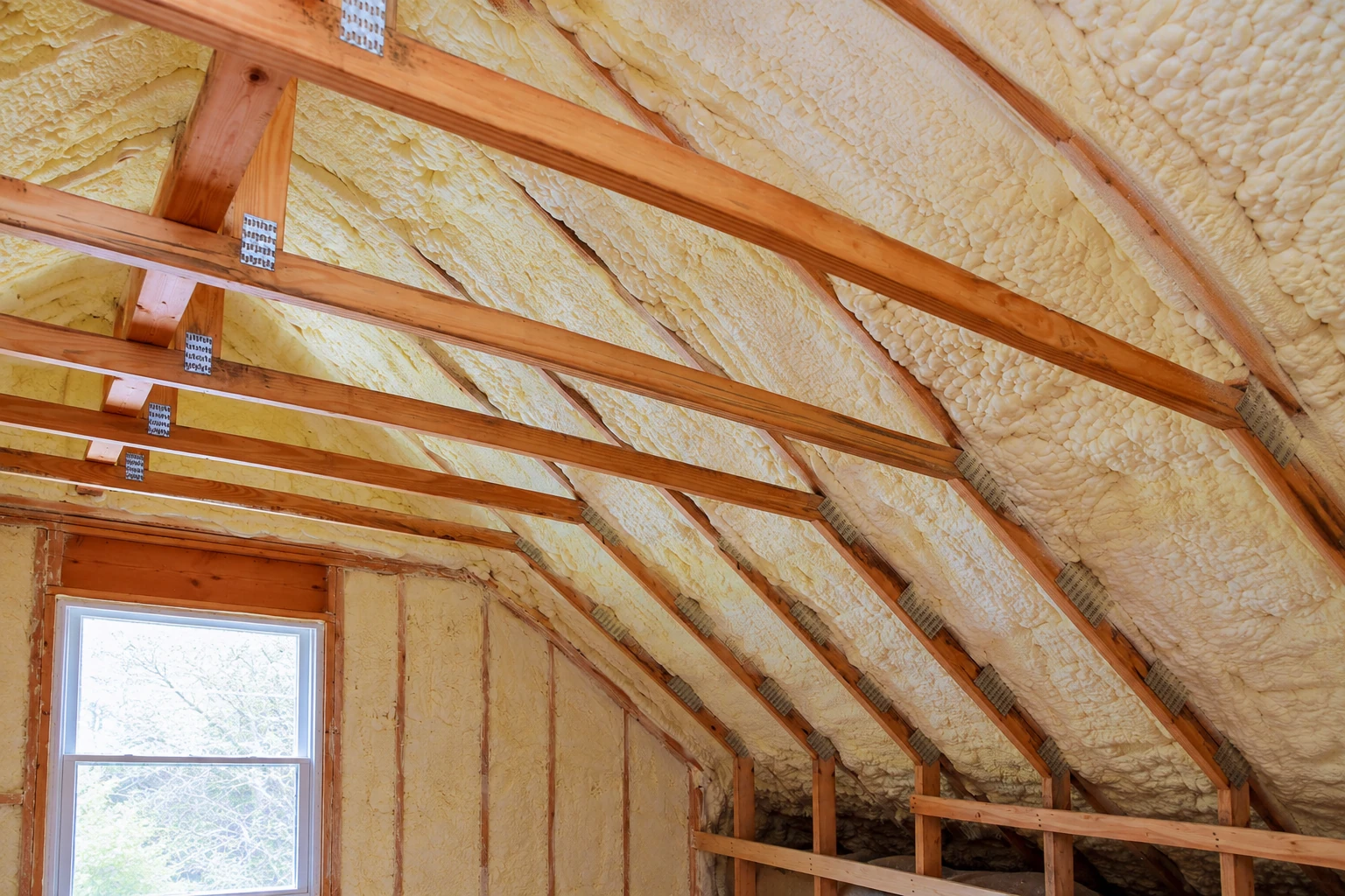 Sloped attic ceiling and walls coated in yellow closed-cell spray foam between exposed wooden rafters.