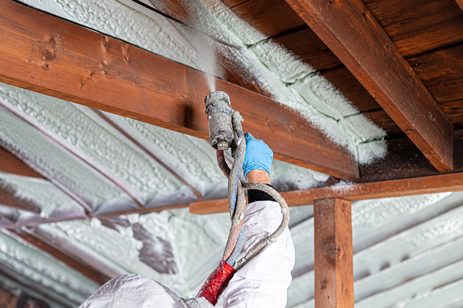 A worker sprays white closed-cell foam insulation between exposed wooden roof beams.