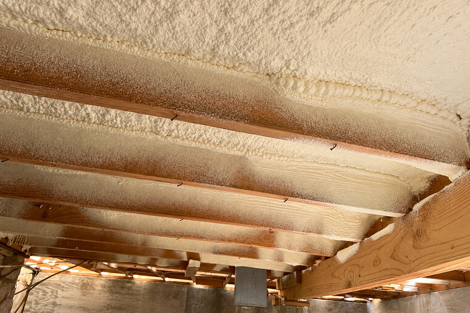 Crawl space ceiling with cream spray foam covering the underside between exposed wooden joists.