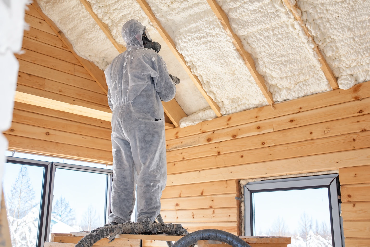 A worker in protective coveralls sprays foam insulation onto a sloped ceiling in a timber-framed room.