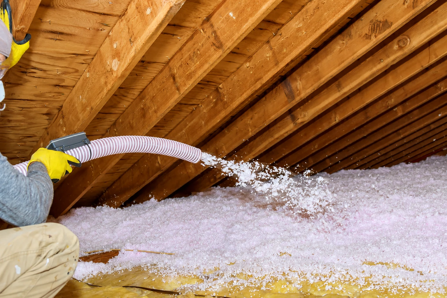 A worker blows pink loose-fill insulation across an attic floor between exposed rafters.